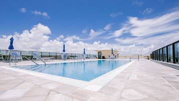 Una piscina con sillas de descanso y sombrillas, rodeada por una terraza de baldosas y una barandilla de vidrio.