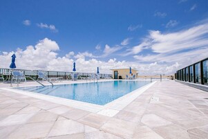 Una piscina con sillas de descanso y sombrillas, rodeada por una terraza de baldosas y una barandilla de vidrio.