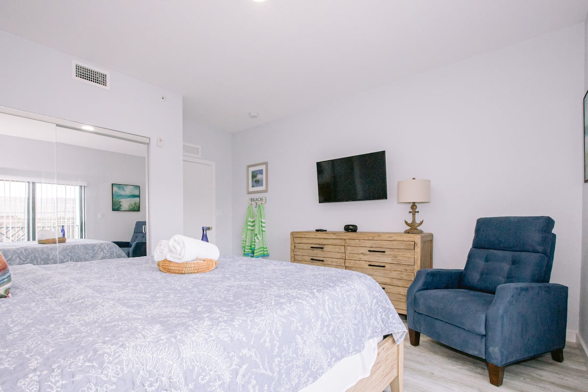 A bedroom with a bed, a dresser, a blue armchair, and a television on the wall.
