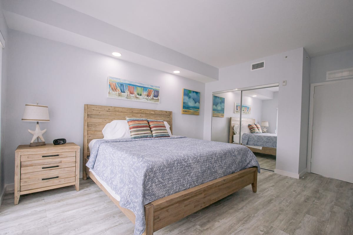 A bedroom with a wooden bed, a nightstand with drawers, a lamp, and a starfish decoration. There are paintings on the wall and a mirror reflecting the room.