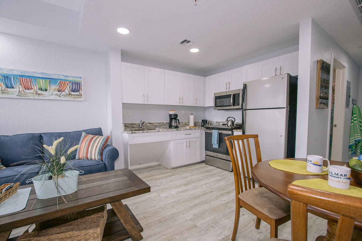 A compact kitchen with white cabinets, a microwave, and a refrigerator. There's a dining table with two chairs and a coffee maker on the counter.