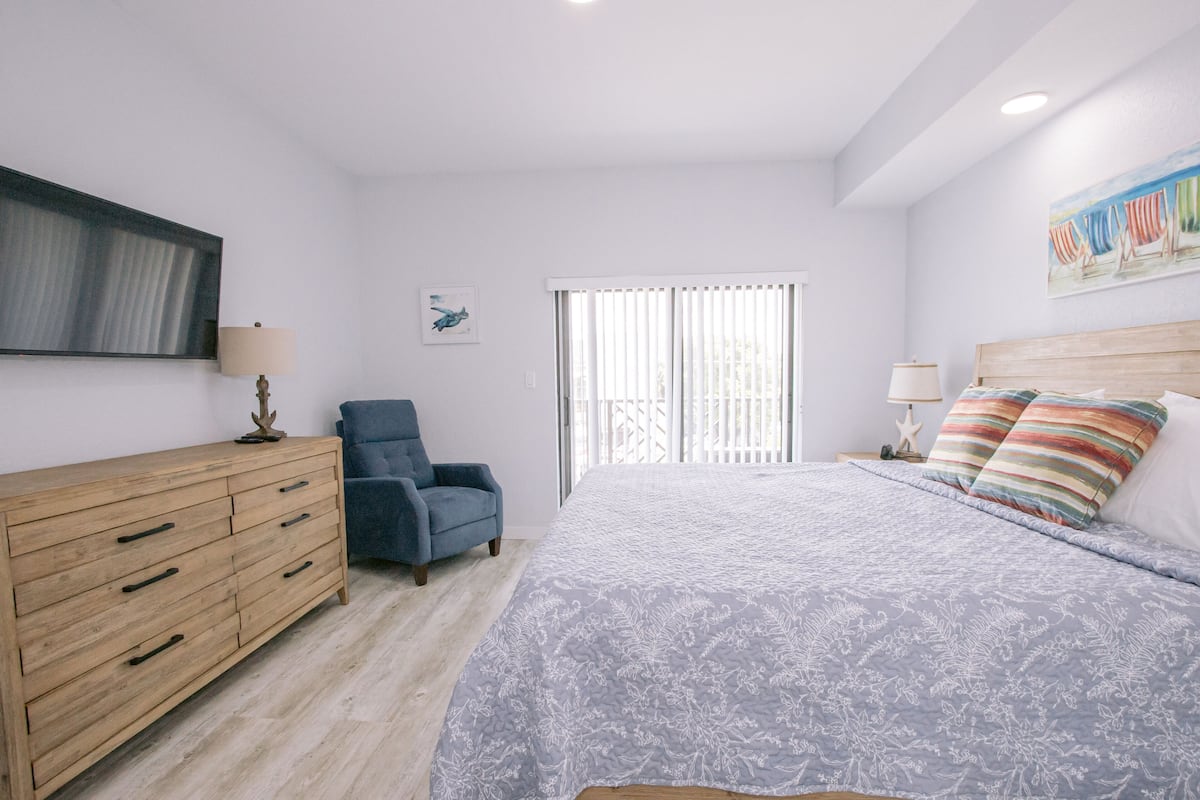 A bedroom with a bed, a dresser, a chair, a window with blinds, and a painting on the wall.