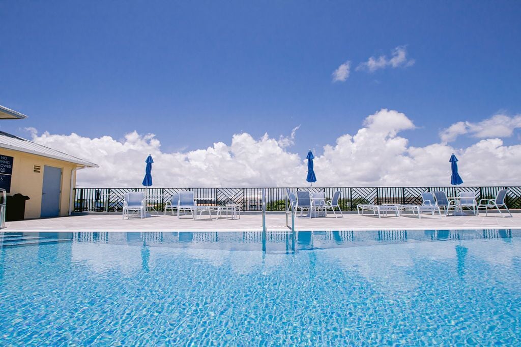 A clear blue pool with lounge chairs and umbrellas, surrounded by a fence and a building with a sign.