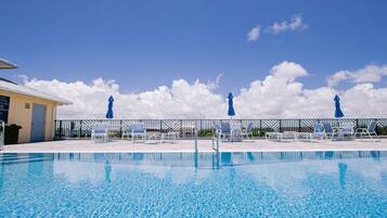 A clear blue pool with lounge chairs and umbrellas, surrounded by a fence and a building with a sign.