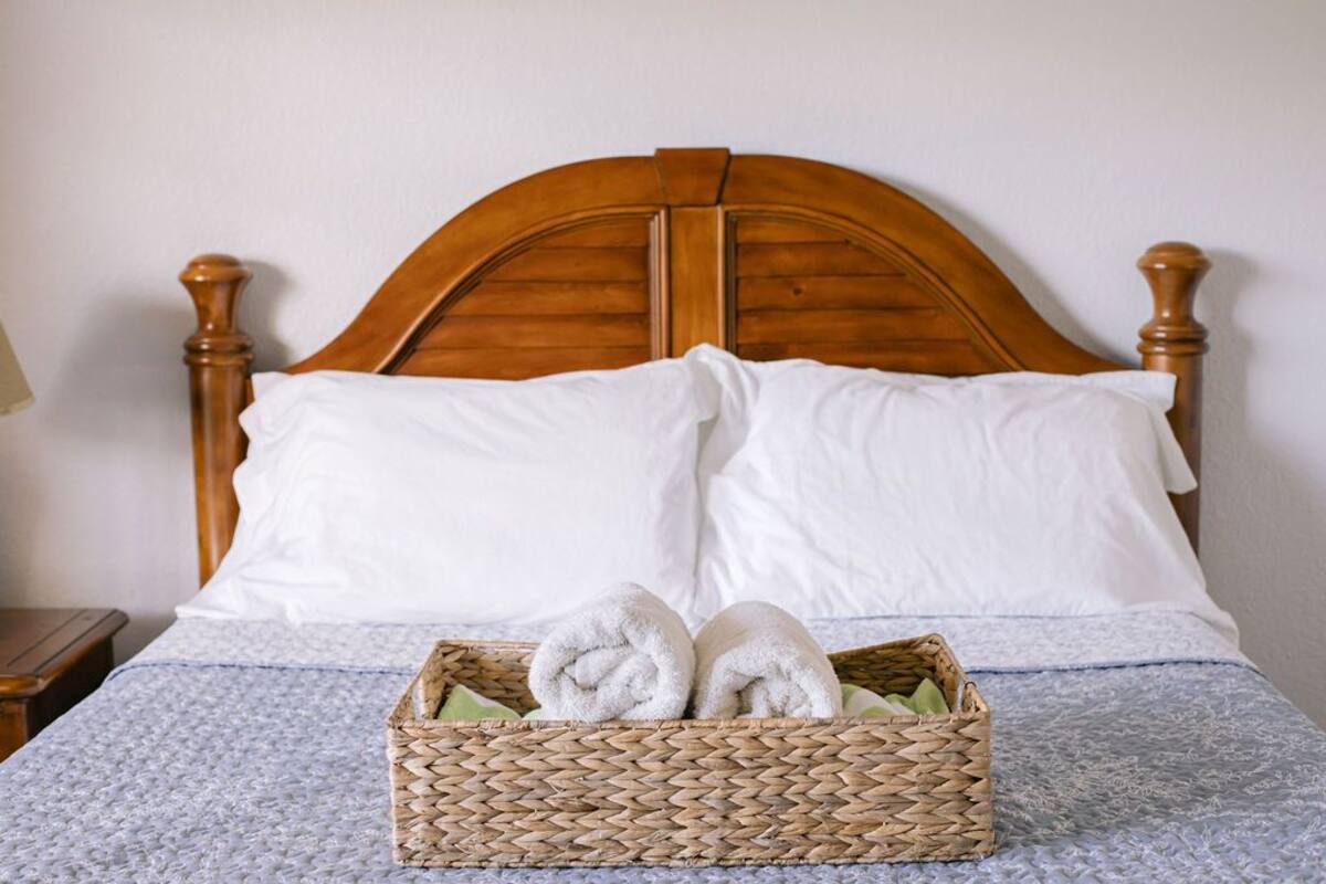 A neatly made bed with a wooden headboard, two white pillows, and a woven basket containing rolled towels.