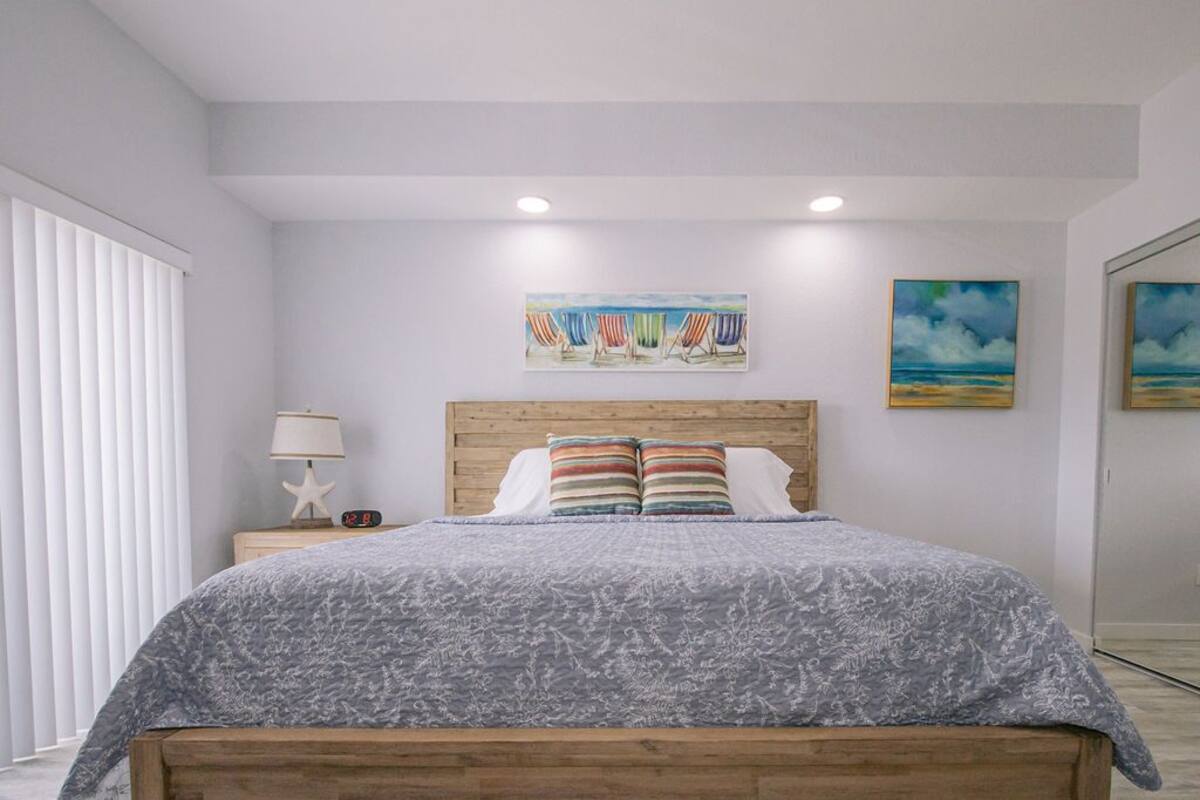 A bedroom with a wooden headboard, a grey patterned bedspread, and two striped pillows. There is a nightstand with a lamp and a starfish decoration. Two paintings are hung on the wall above the bed.