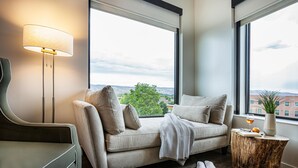 A modern living room with a sofa, a side table, a lamp, and a view of the city through large windows. - Hotel Maverick (Grand Junction)