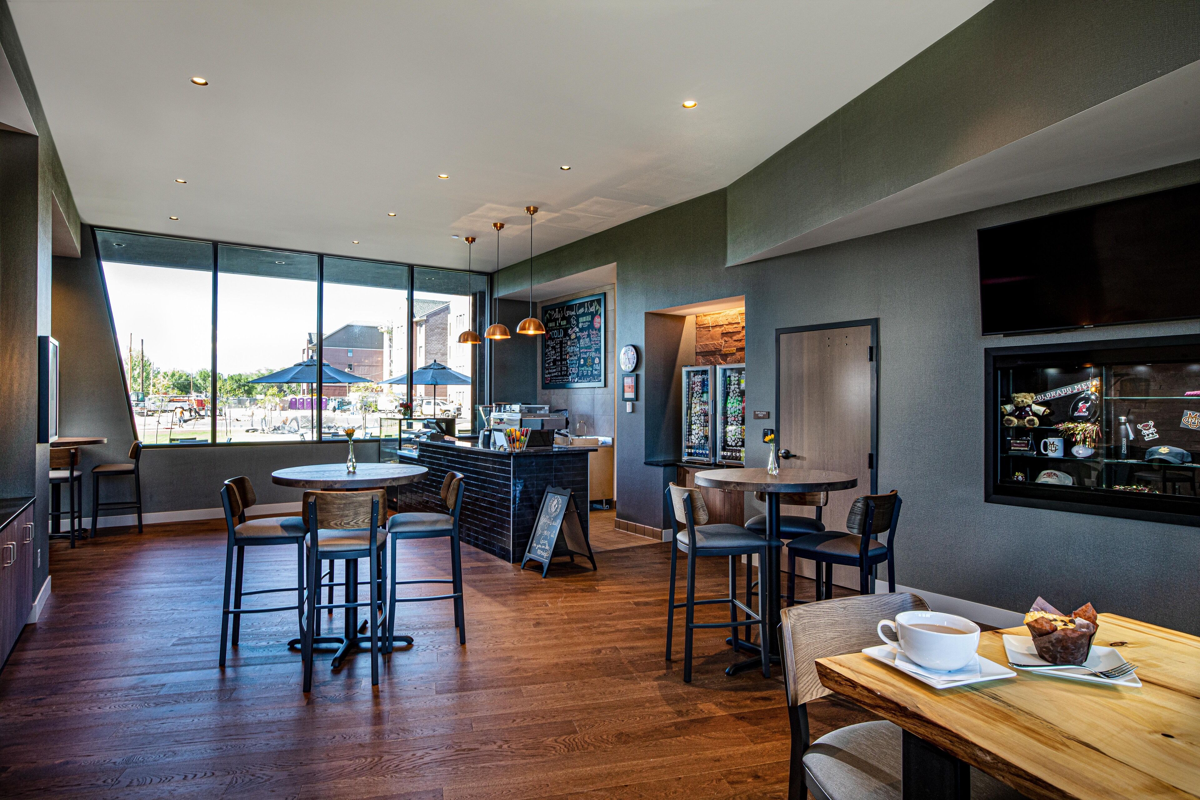 A modern cafe with wooden floors, multiple seating areas, a bar, and large windows offering a view of the outside.