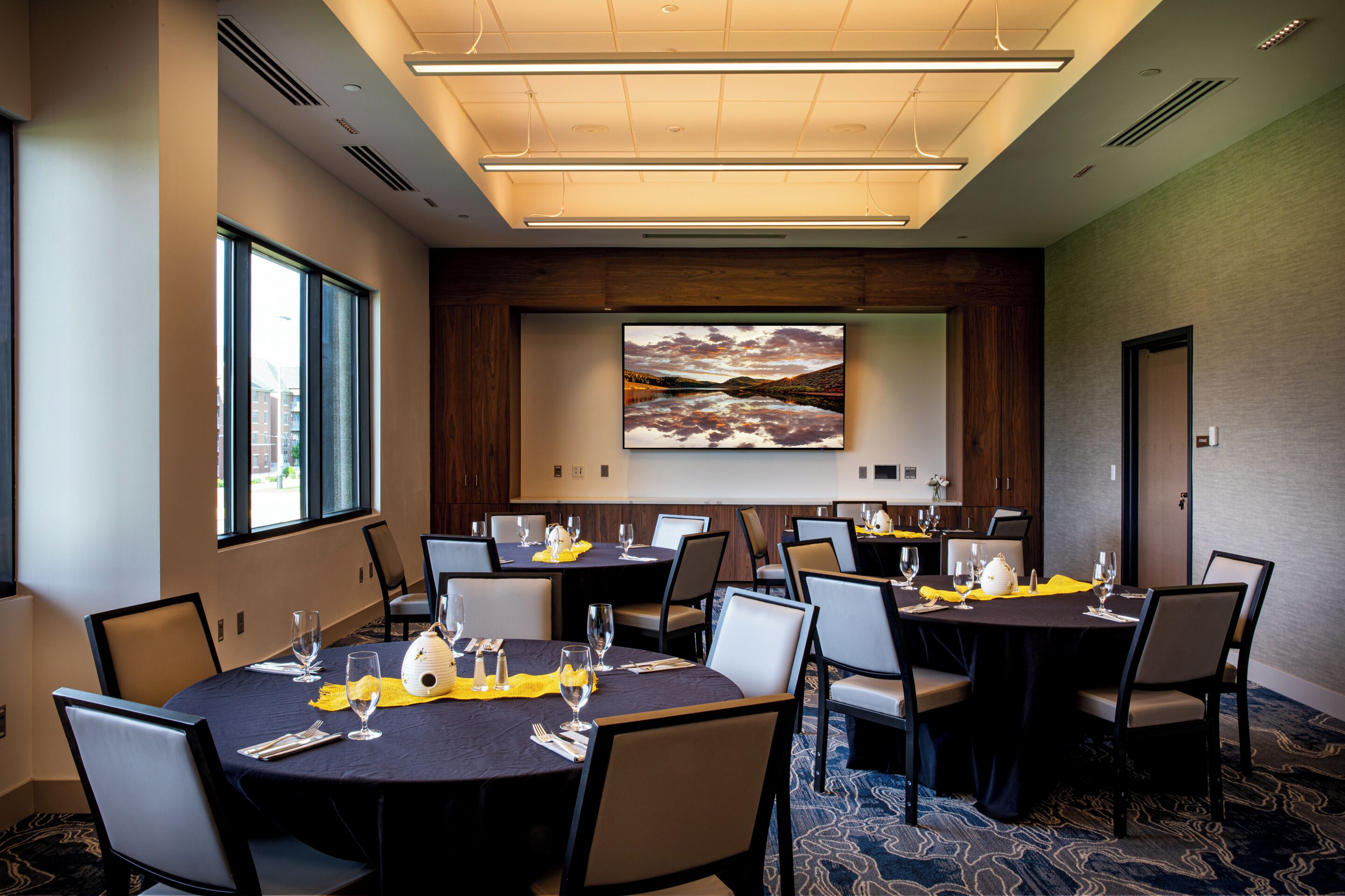A conference room with multiple tables and chairs, equipped with glassware and napkins, and a large screen displaying a scenic image.