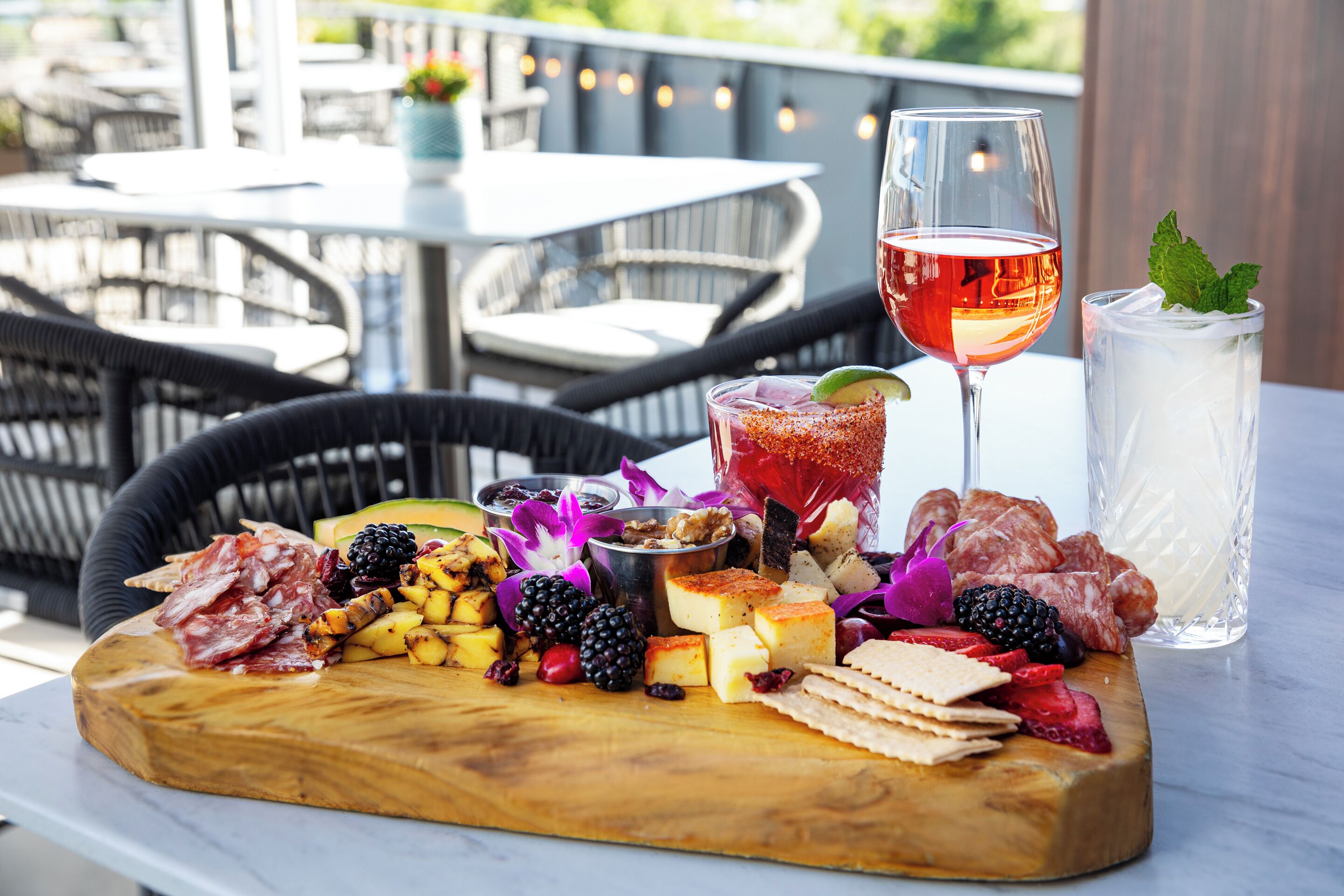 A charcuterie board with various cheeses, meats, fruits, and nuts on a wooden board, accompanied by a glass of red wine and a glass of iced beverage on a table.