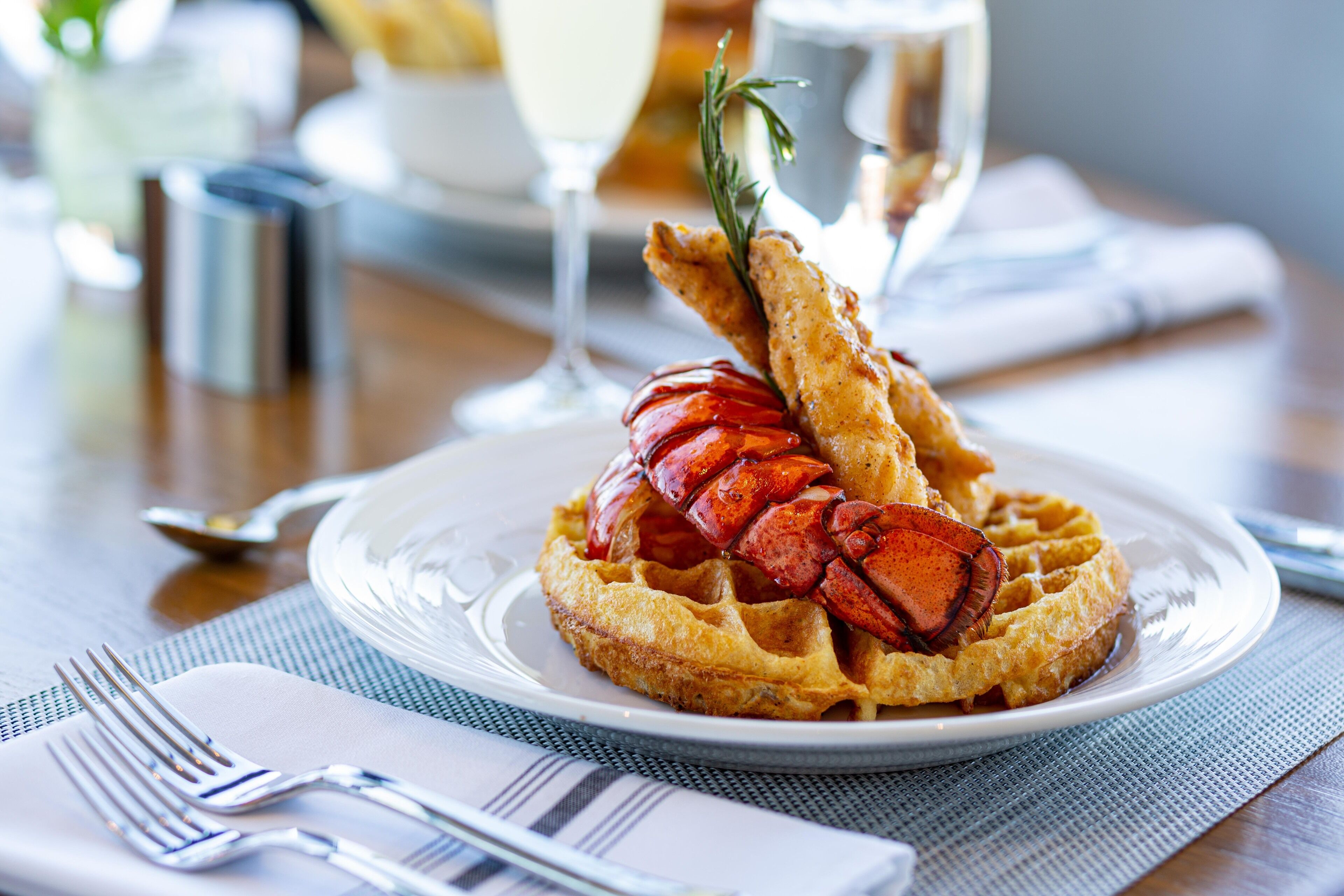 A plate with a lobster tail on a waffle, a glass of white wine, and a fork on a napkin.