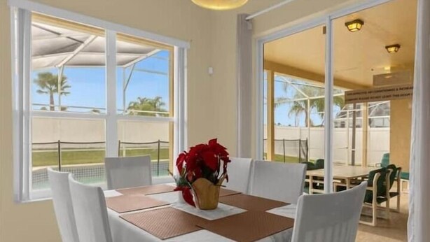 A dining area with a table set for two, a vase of red flowers, and large windows offering a view of a pool and palm trees. - Fun Family Escape: 6 BR Oasis w/Private South-Facing Pool! (Davenport)