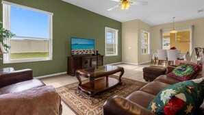 A living room with a brown leather sofa, a wooden coffee table, and a television on a stand. There is a ceiling fan, a window with a view of the outdoors, and a dining area in the background. - Fun Family Escape: 6 BR Oasis w/Private South-Facing Pool! (Davenport)