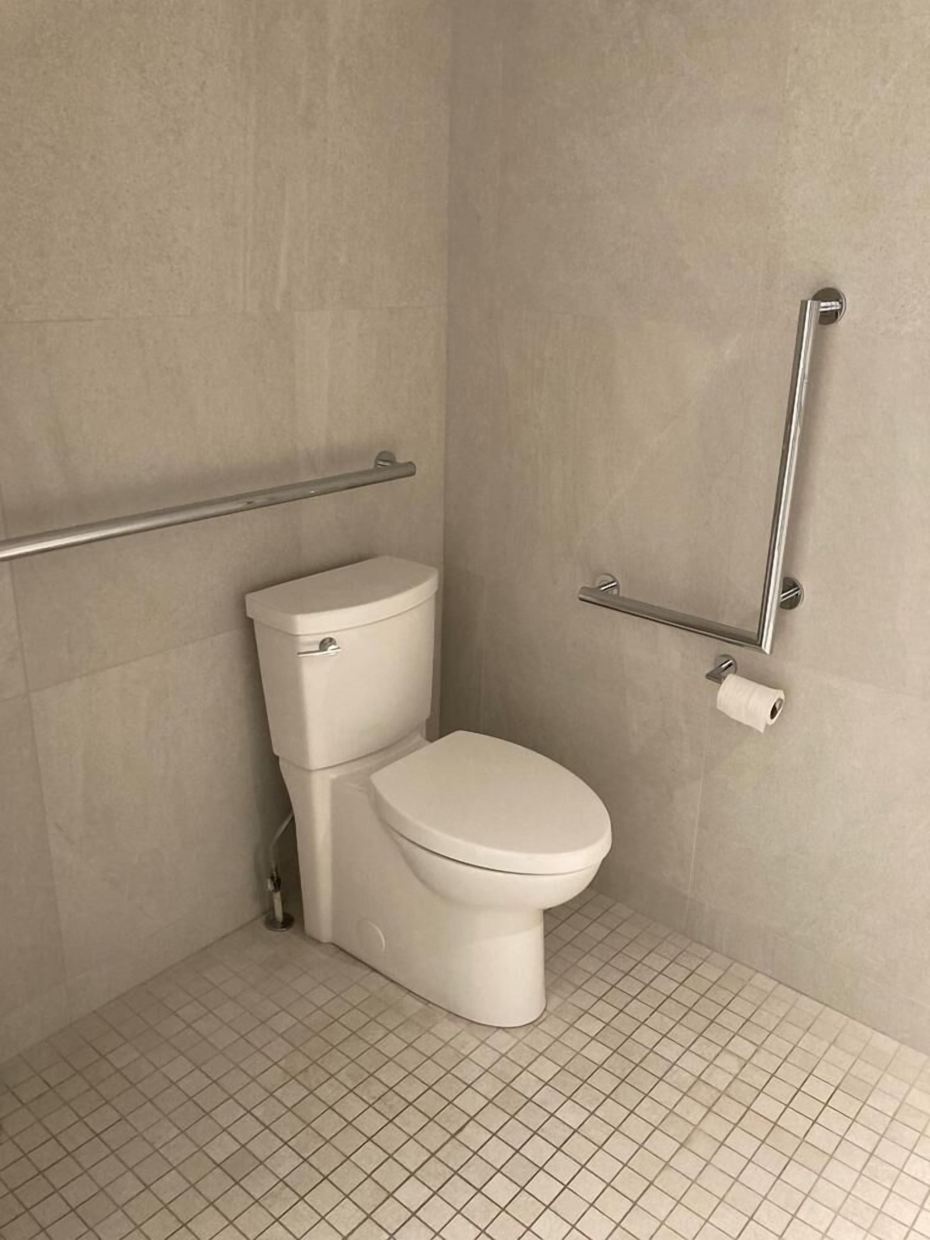 Um banheiro pequeno, revestido com azulejos, com vaso sanitário, barras de apoio e porta-papel higiênico.
