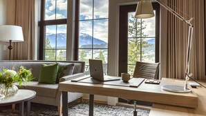 A modern hotel room with a large window offering a mountain view, a desk with a laptop, a lamp, and a coffee cup. - Headwaters Lodge at Eagle Ranch Resort (Invermere)