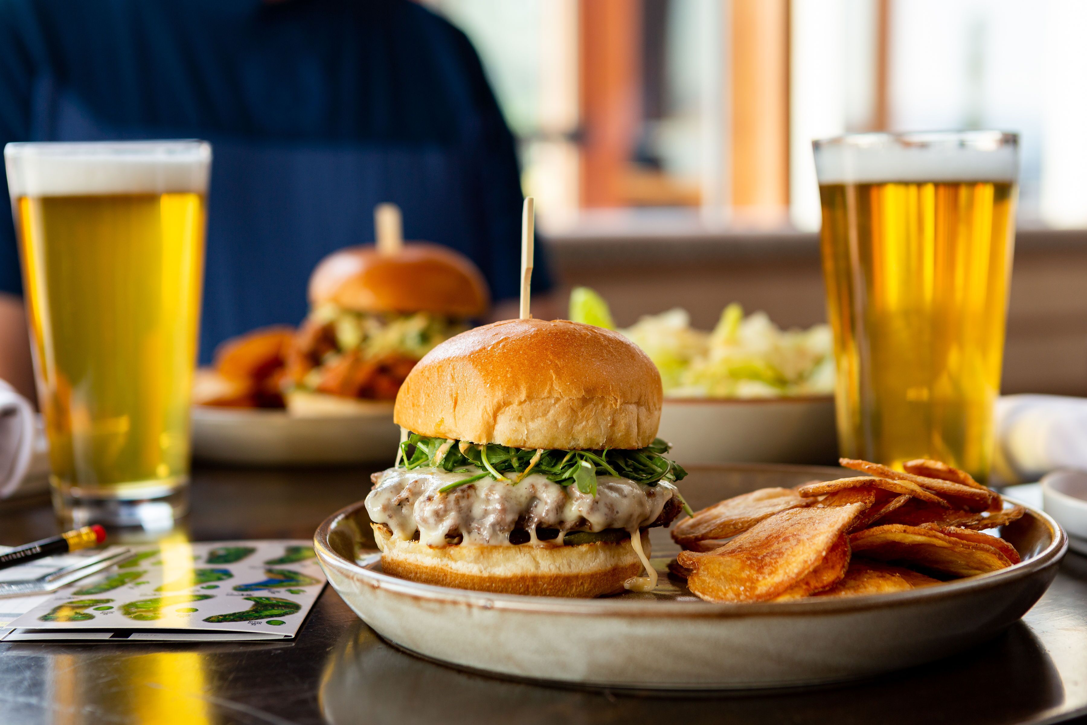 Un hamburger avec un cure-dent, une assiette de frites et deux verres de bière sur une table.