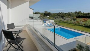 A balcony with a glass railing overlooking a swimming pool, a dining table with chairs, and a view of a landscaped garden.