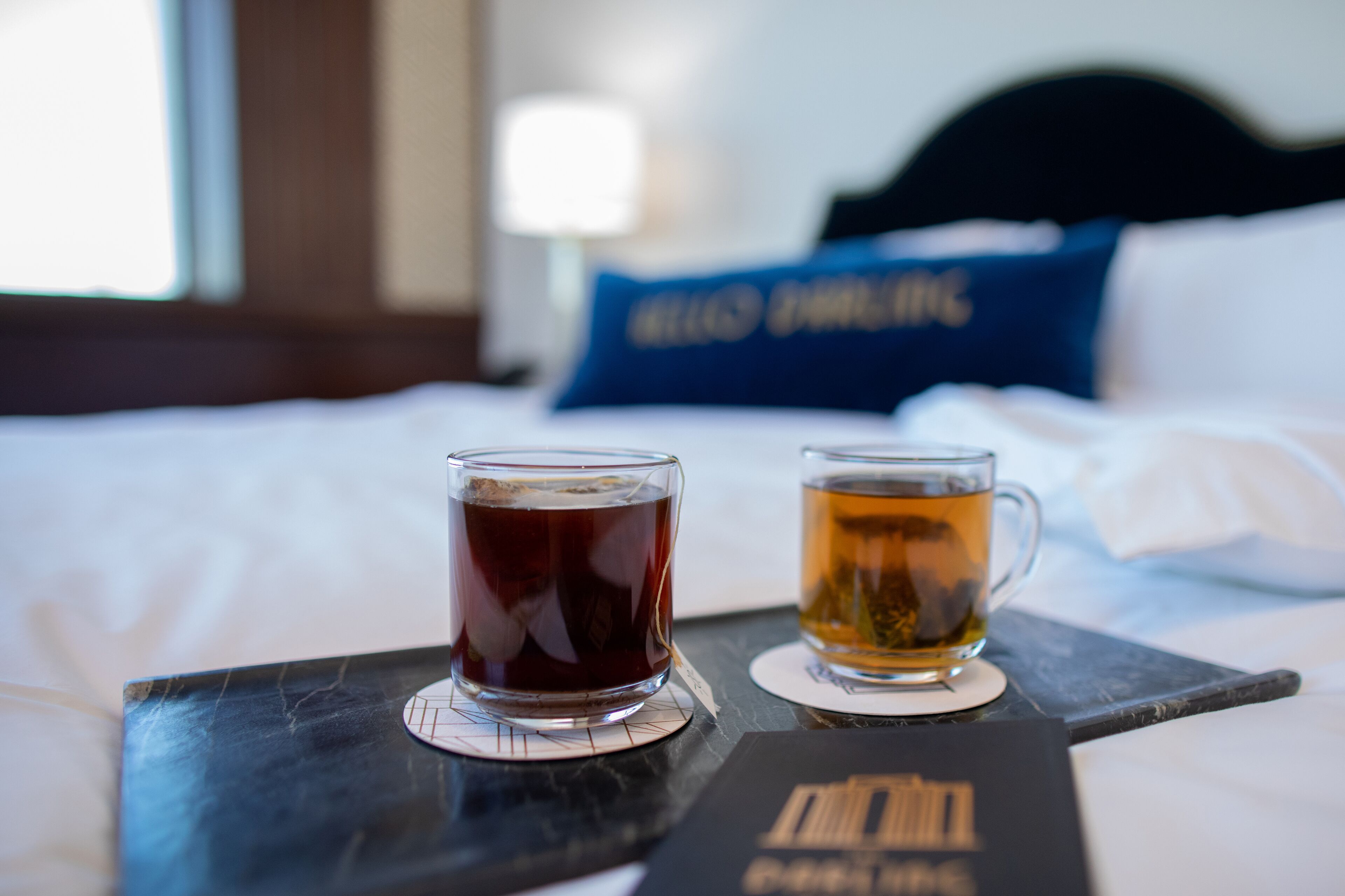 A tray with two glasses of tea on a bed with white sheets and a blue pillow in the background.