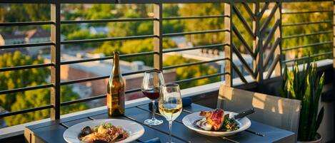 Un tavolo imbandito con piatti di cibo, bicchieri da vino e una bottiglia, su un balcone affacciato su vegetazione e edifici.