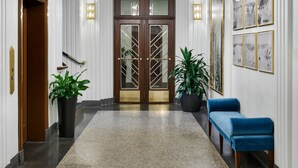 A hotel lobby with a wooden double door, a marble floor, potted plants, a blue bench, and framed artwork on the walls.