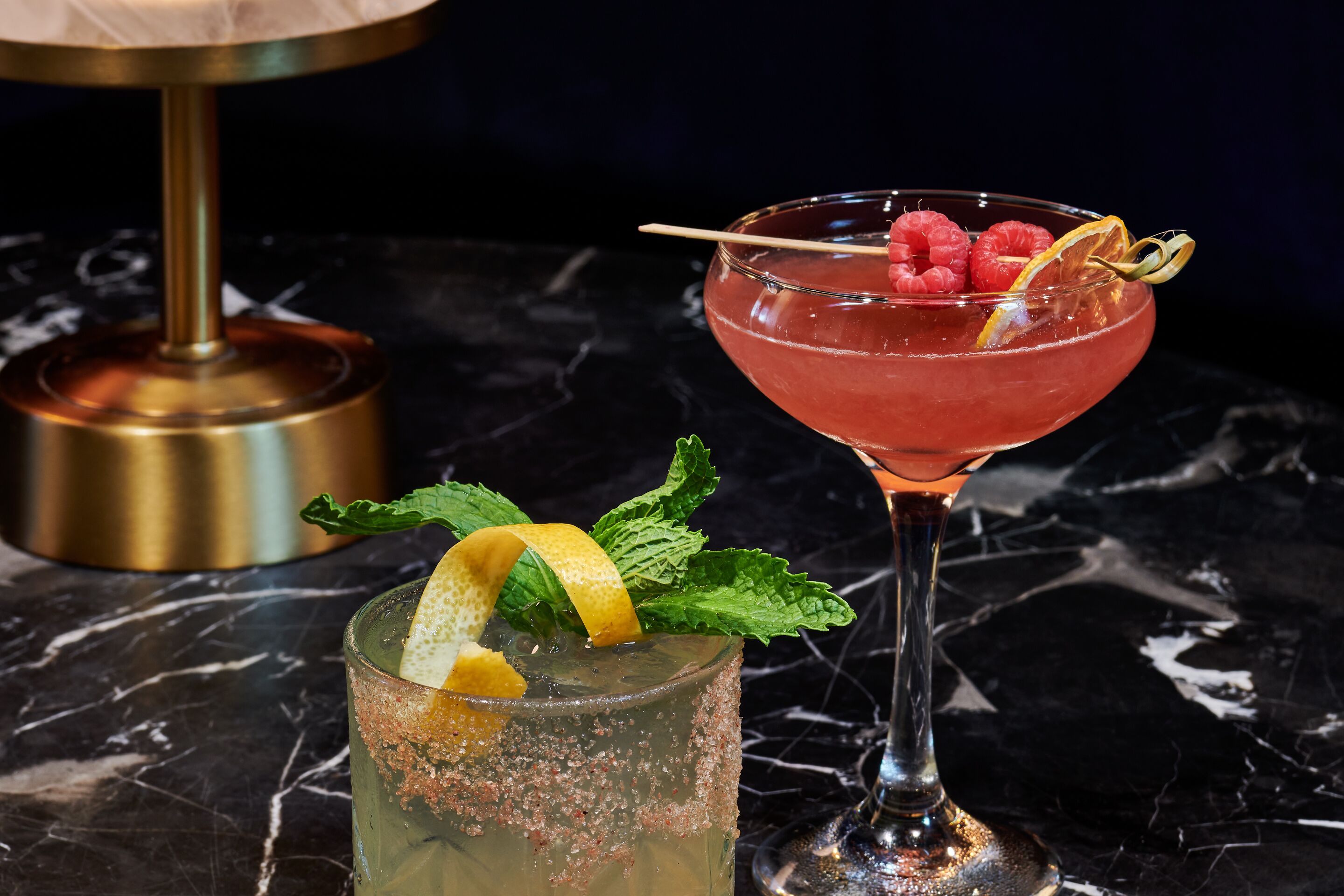 Two cocktails on a marble table with a gold base and a dark background.