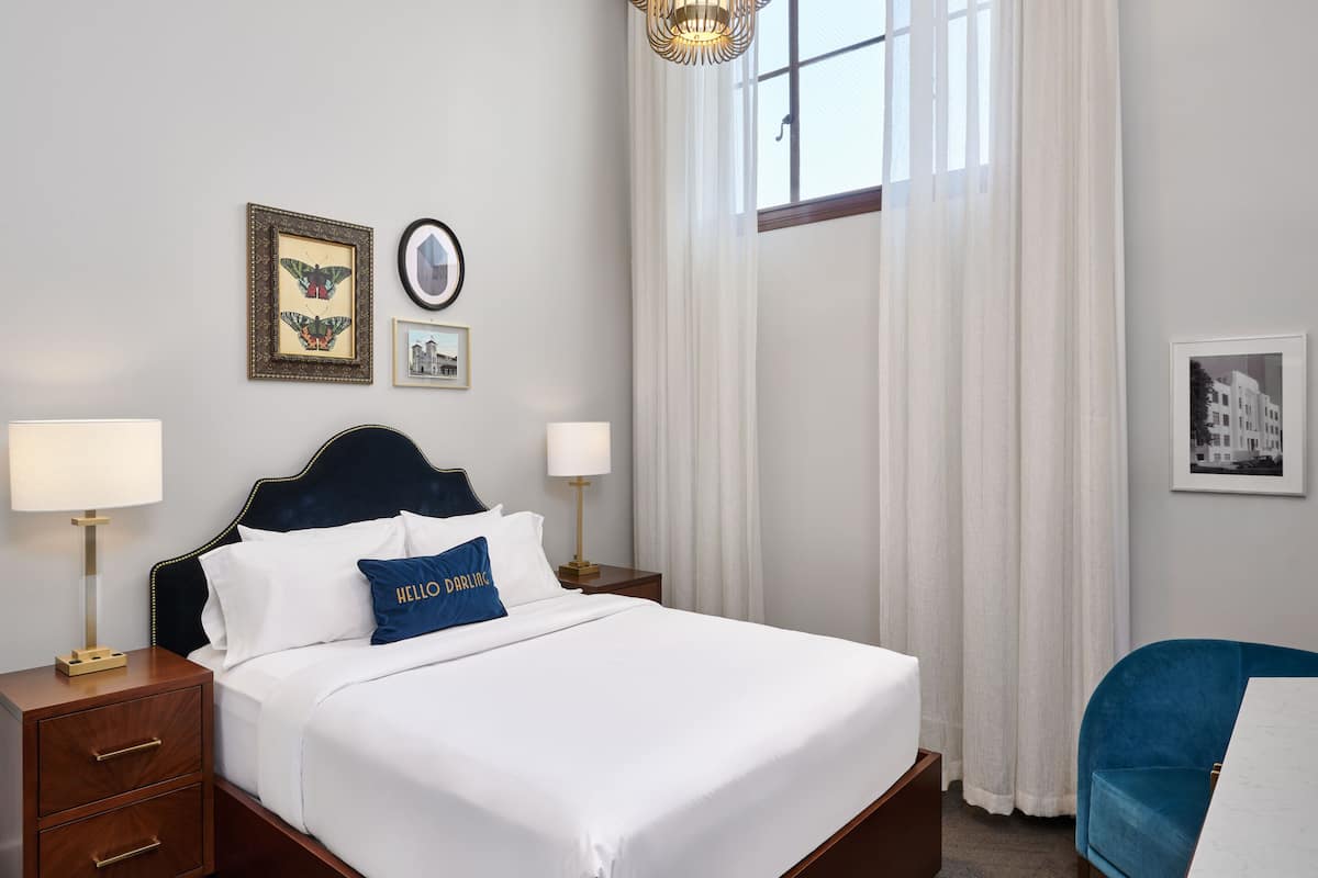A neatly made bed with white linens and a blue pillow, a bedside table with a lamp, and a blue chair.