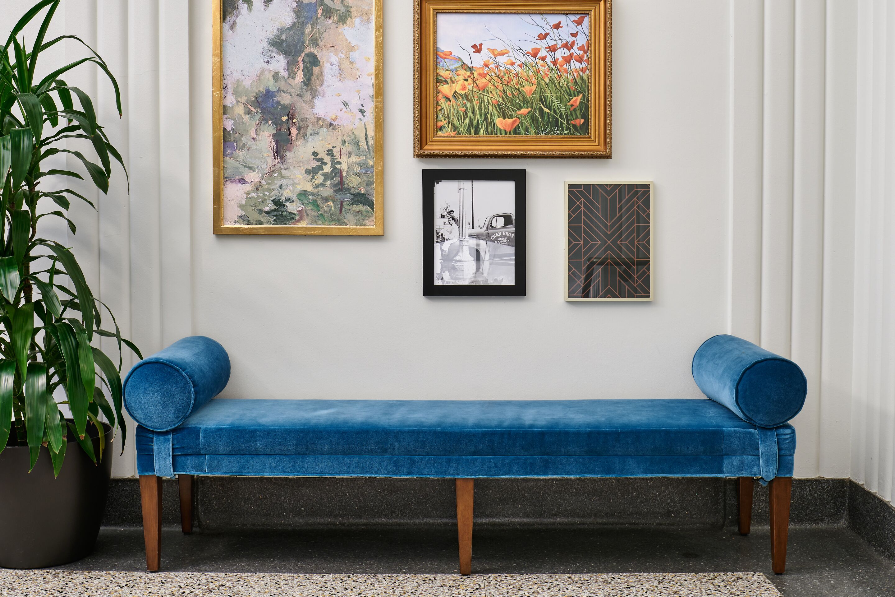 A blue bench with wooden legs, a potted plant, and three framed artworks on a white wall.