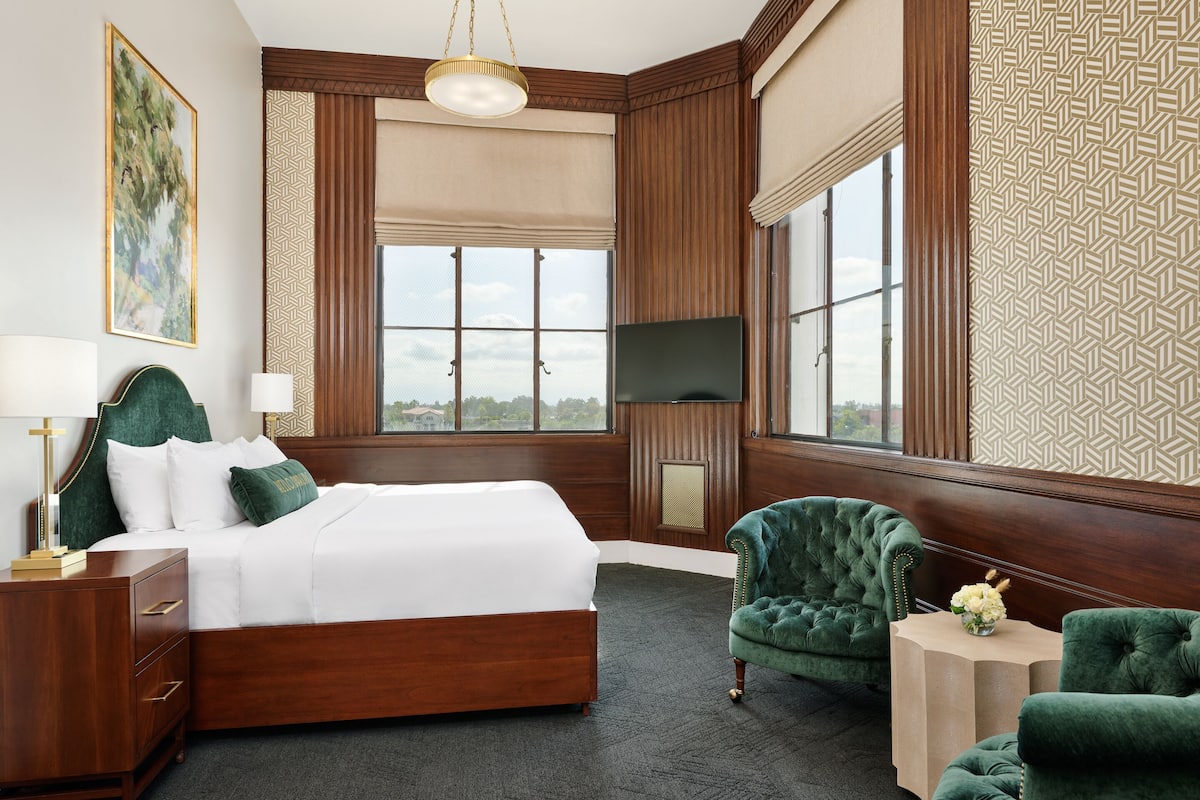 A hotel room with a large bed, a TV, a green armchair, and a view of the city.