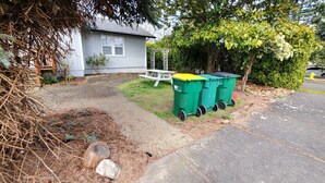 Zona residencial com uma casa, uma mesa de piquenique e dois contentores verdes para lixo.
