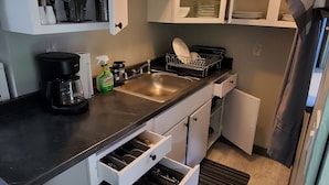 Cozinha compacta com armários brancos, bancada preta e pia de aço inoxidável. Há utensílios e uma cafeteira na bancada.