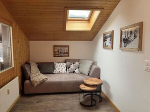 A cozy room with a wooden ceiling, a skylight, a sofa with cushions, a small wooden table, and two framed pictures on the wall.