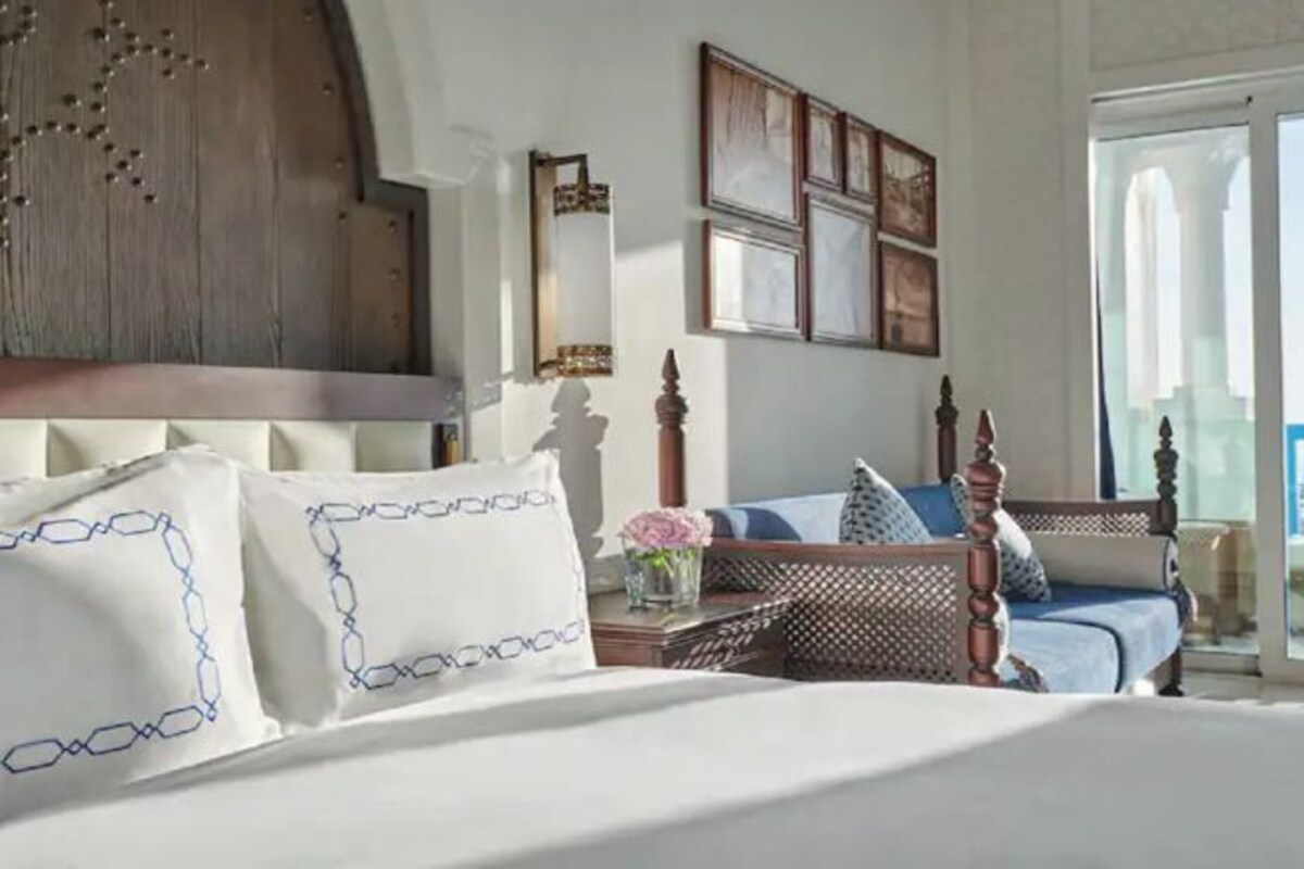 Une chambre avec un grand lit, une tête de lit en bois, une table de chevet ornée d’un vase de fleurs et une fenêtre donnant sur un beau paysage.