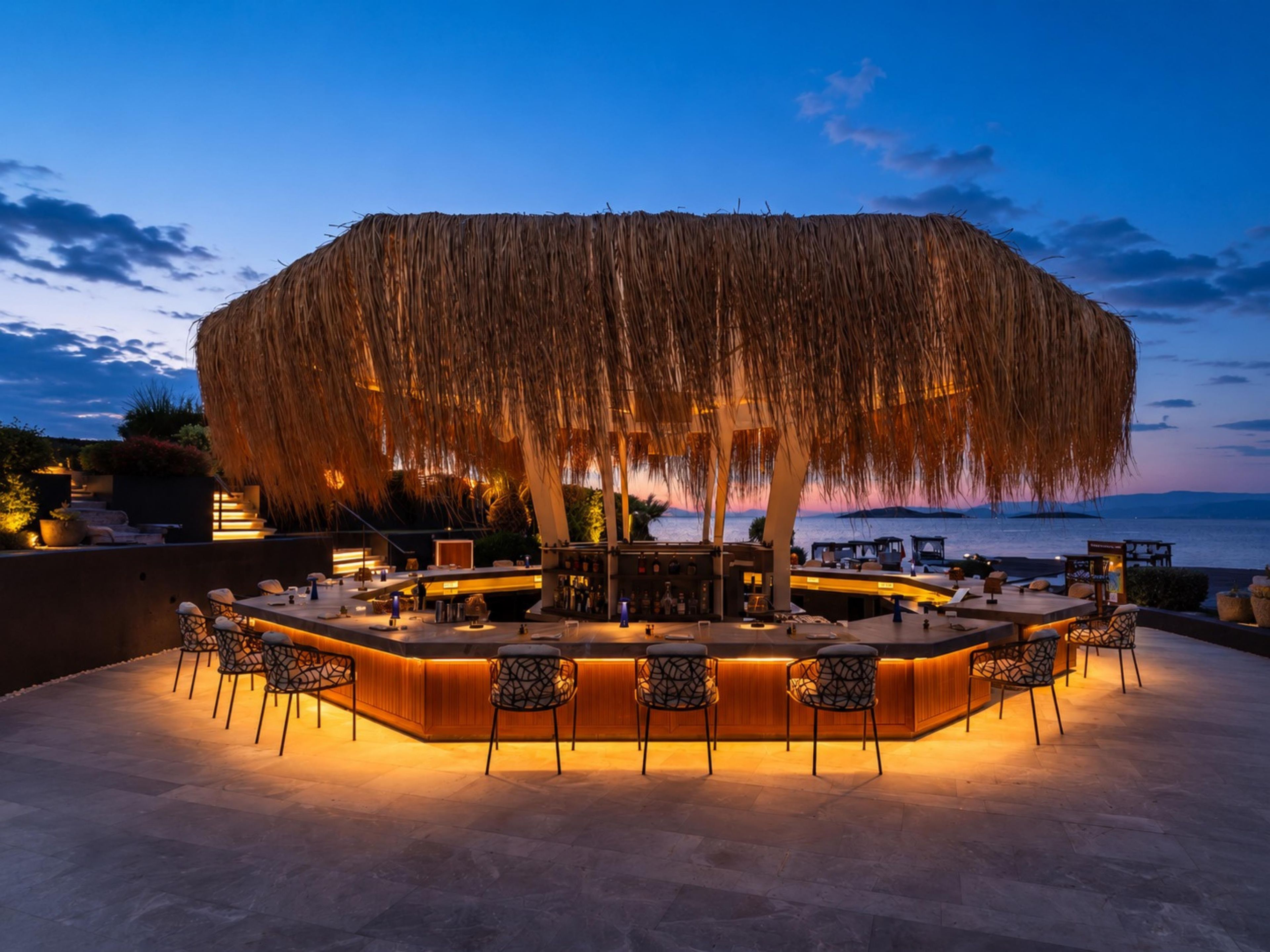 Zona de bar al aire libre con techo de paja, asientos y vista al mar.