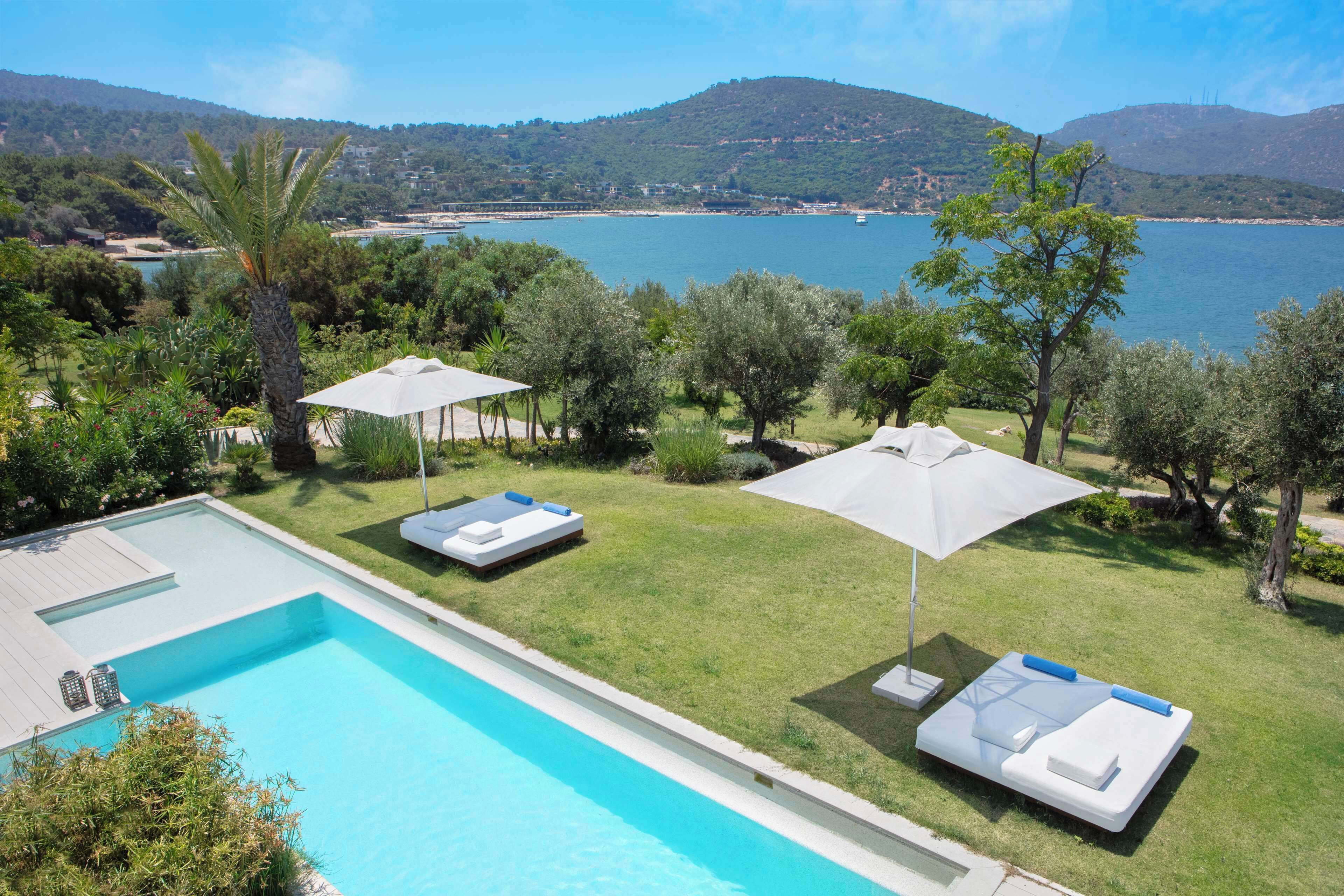 A swimming pool with lounge chairs and umbrellas, surrounded by greenery and a view of the sea and hills.