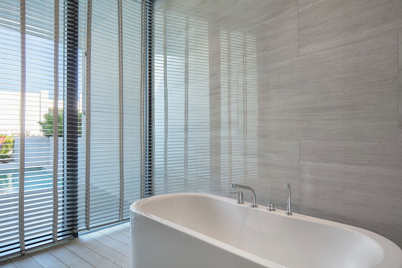 A modern bathroom with a freestanding bathtub, large windows with blinds, and a tiled wall.