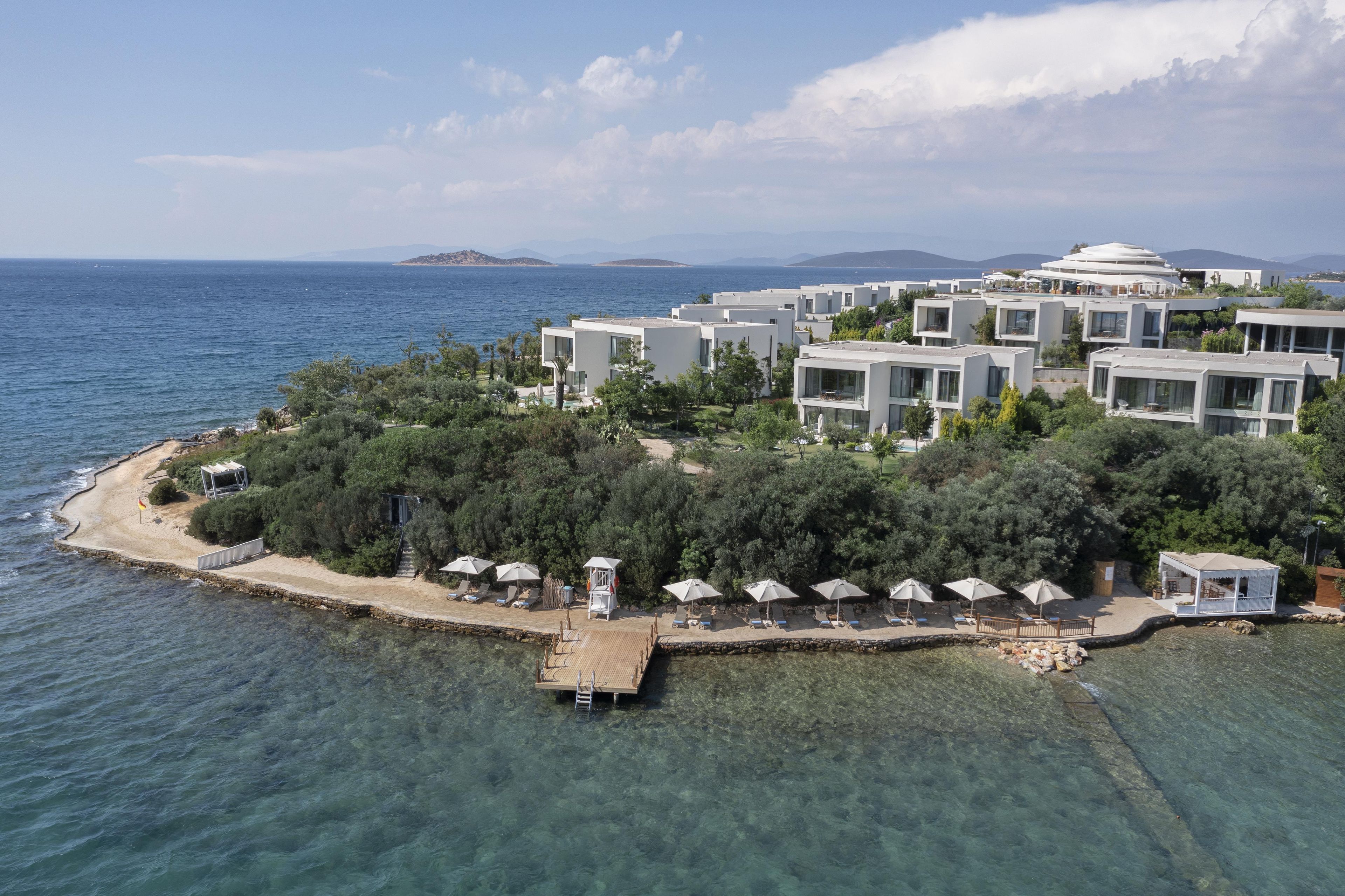 A coastal resort with modern buildings, clear blue sea, and a wooden dock with umbrellas.