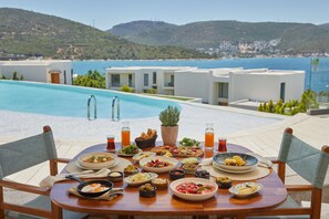 Une table dressée avec différents plats et boissons, donnant sur une piscine et offrant une vue panoramique sur des collines et la mer.