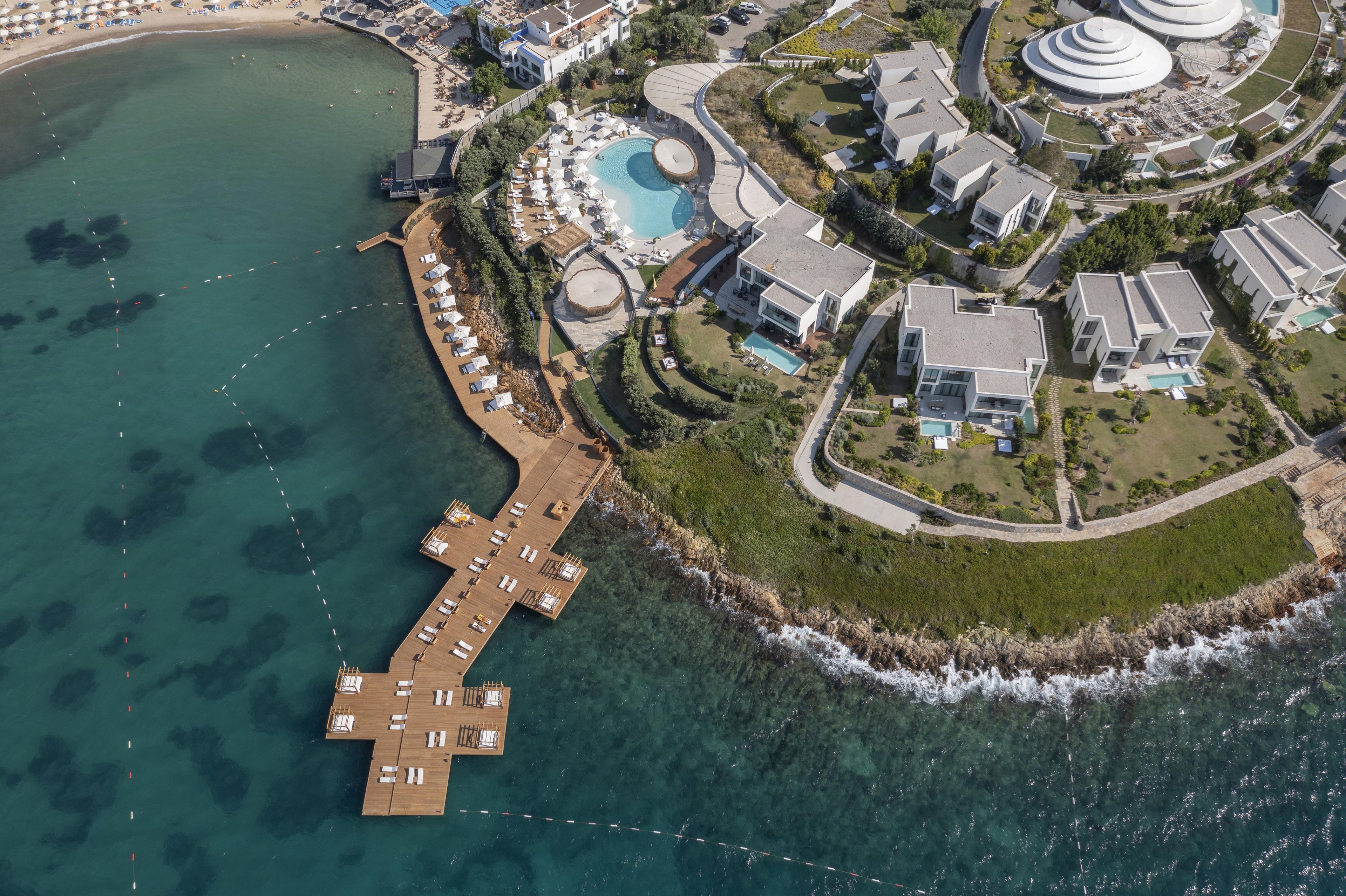 Vista aérea de un complejo turístico costero con un muelle de madera, una piscina y edificios modernos.
