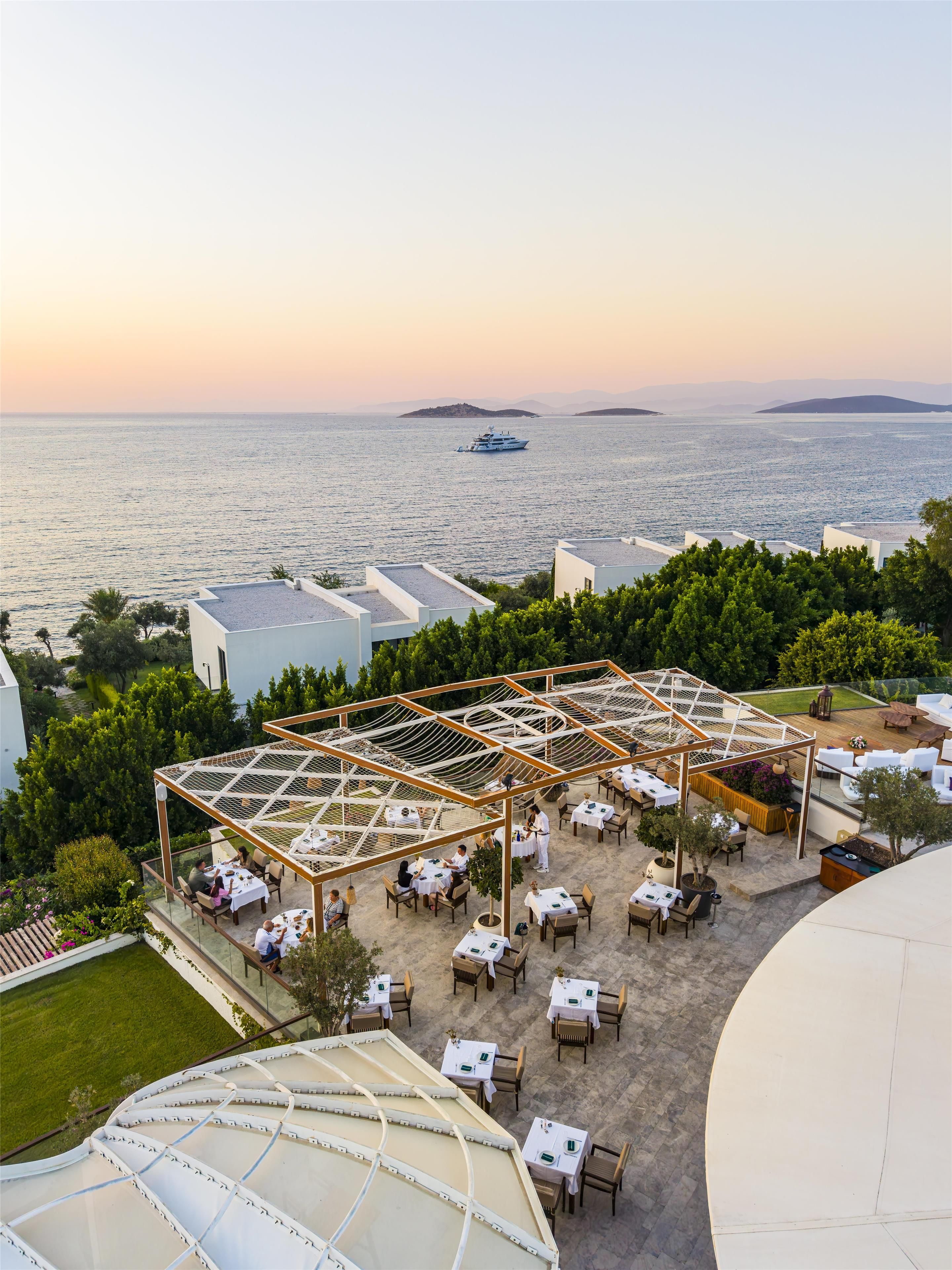 Un espacio al aire libre con mesas y sillas, rodeado de vegetación y vistas al mar.
