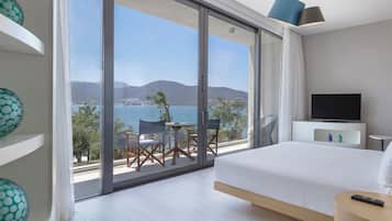 Quarto de hotel com uma cama grande, televisão, varanda com mesa e cadeiras, e vasos decorativos.