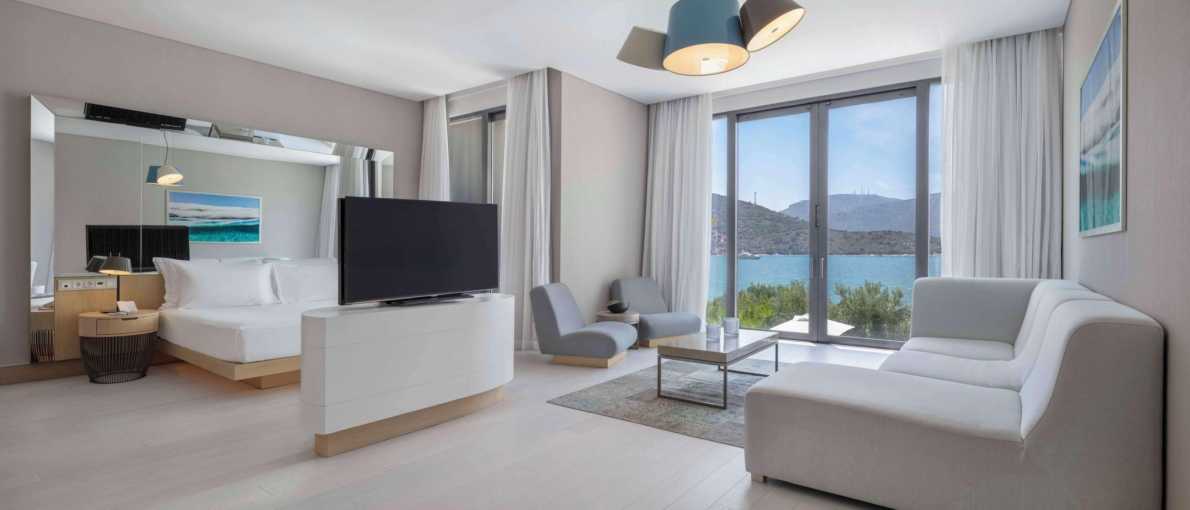 A modern hotel room with a large bed, a TV, a sofa, and a dining area with a view of the mountains.