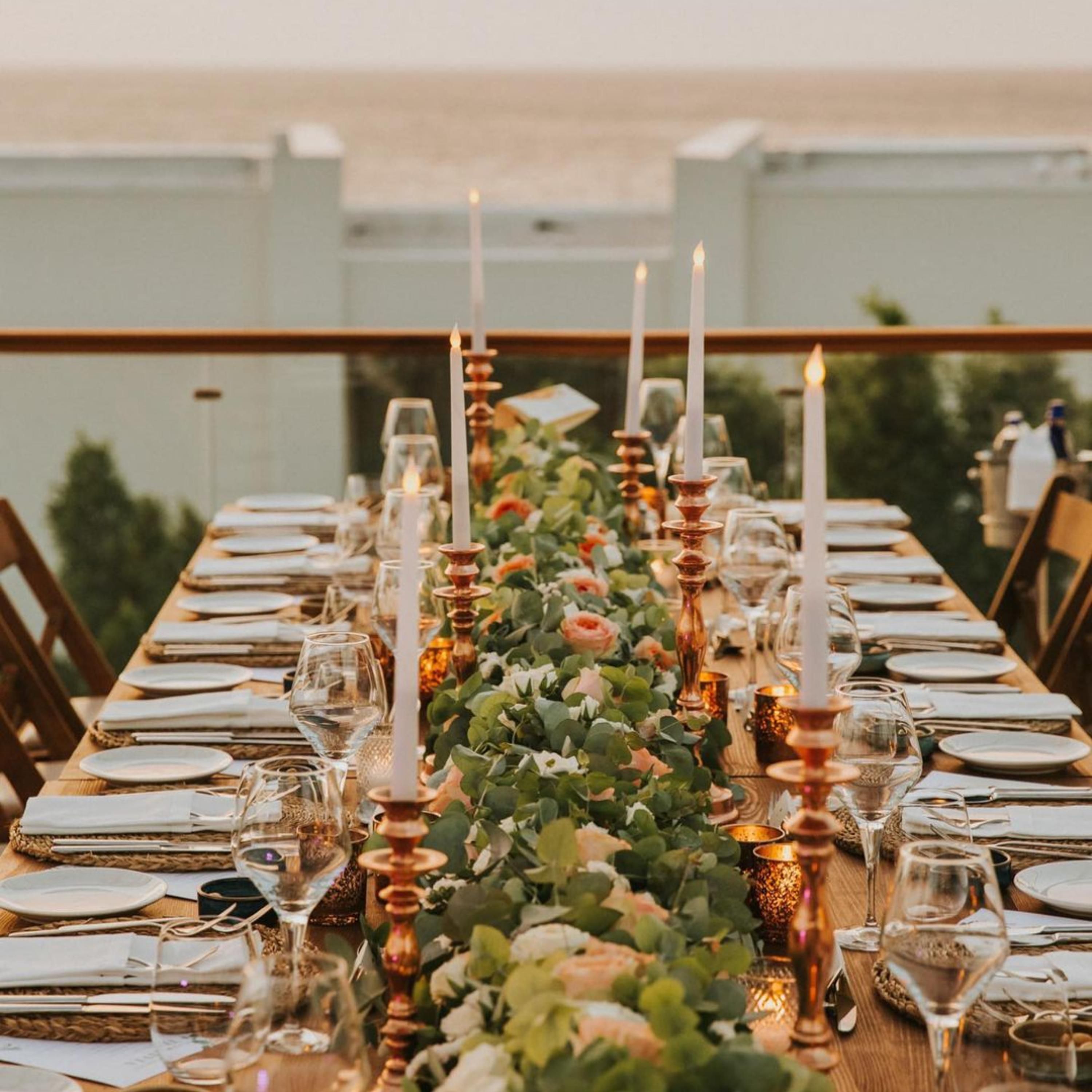 Una larga mesa de comedor dispuesta con velas, cristalería y un centro de mesa floral.