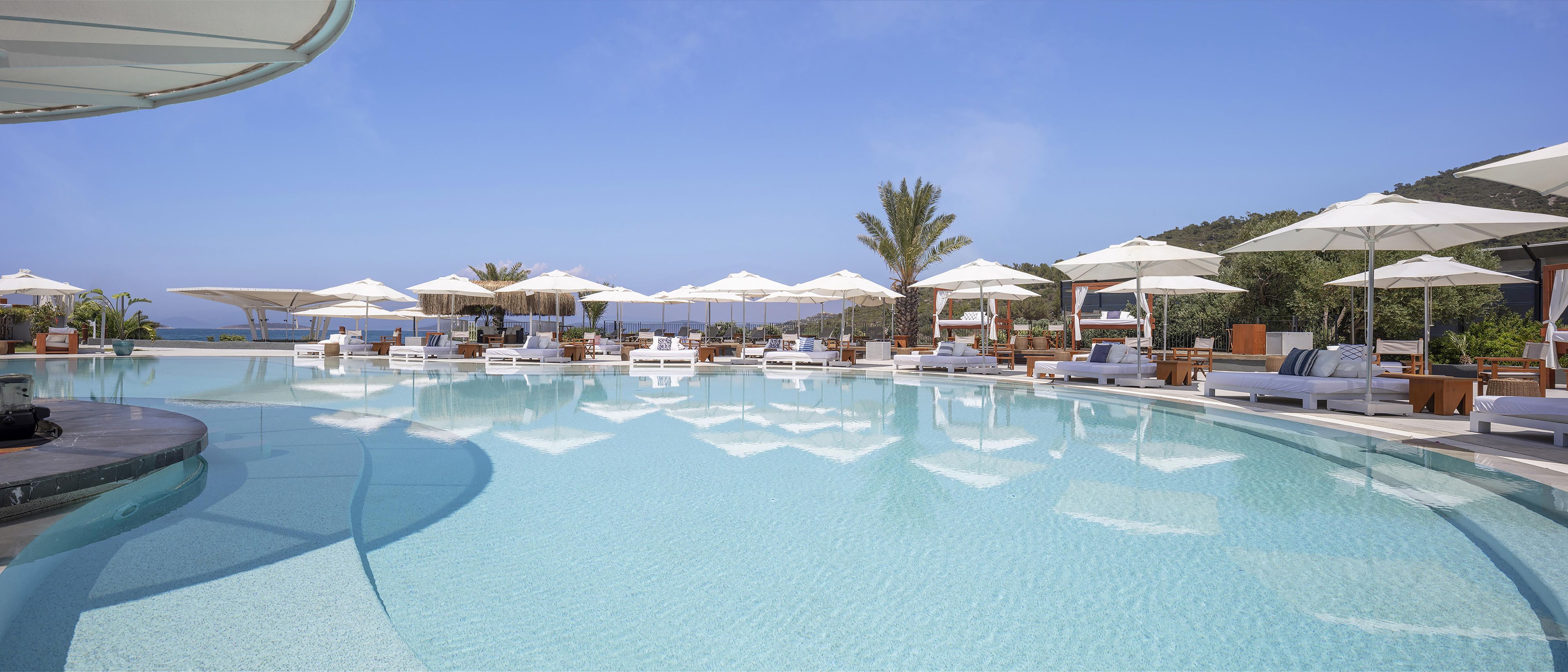 A poolside area with sun loungers and umbrellas, a clear sky, and a view of the sea.
