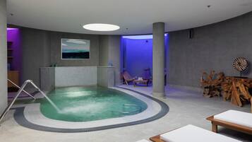 A modern indoor pool area with a clear water feature, lounge chairs, and a wooden sculpture.