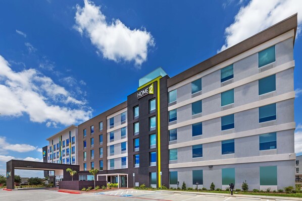 Ett hotell med flera våningar och modern design, med en tydlig skylt som visar ”HOME2”, stora fönster och en parkeringsplats med markerade parkeringsrutor.