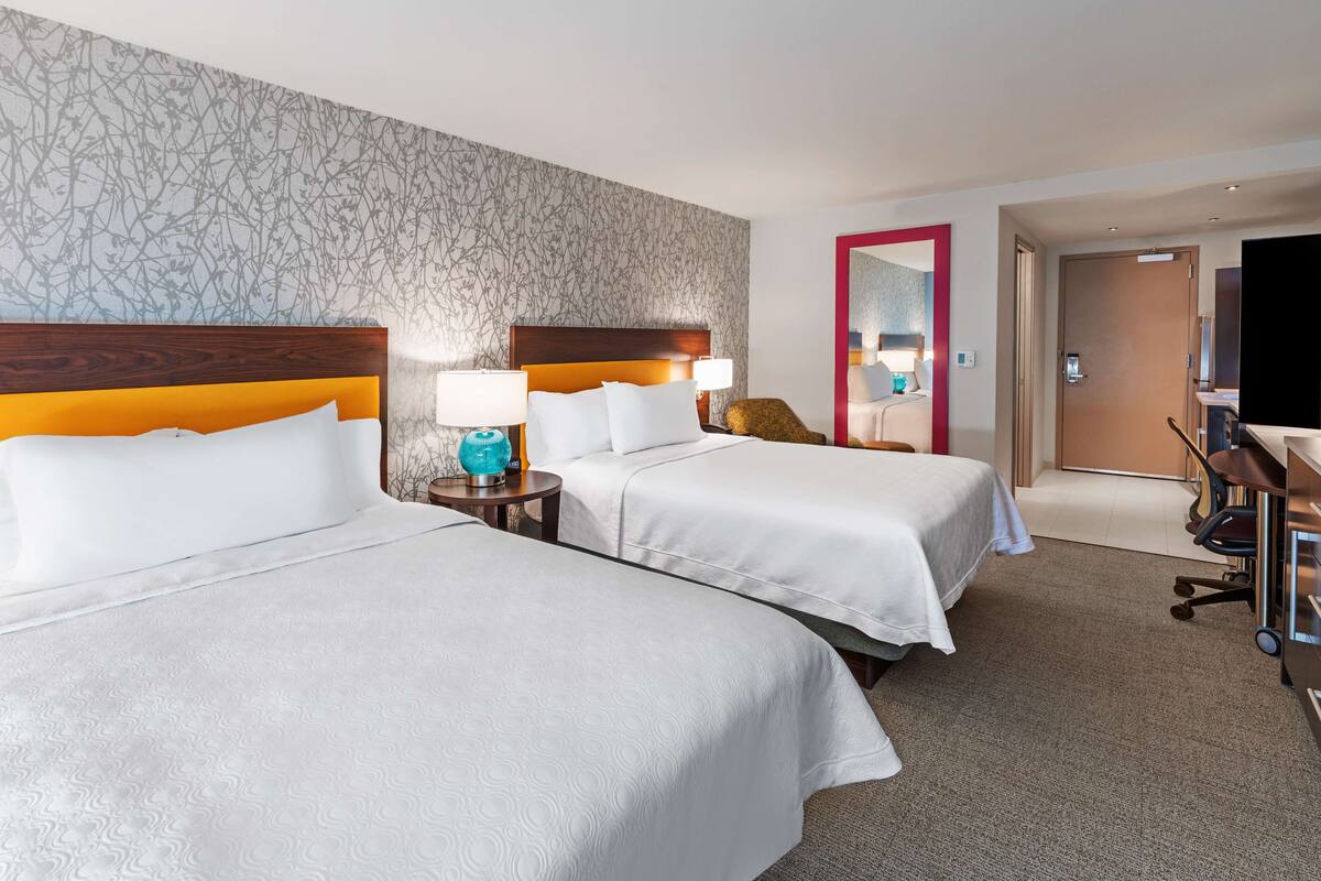 Une chambre d’hôtel avec deux lits, un miroir, un bureau avec une chaise et une porte donnant sur une autre pièce.