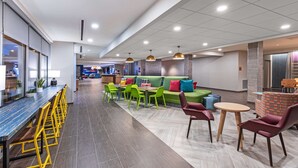 Ein moderner Loungebereich mit einer langen Bar, grünen Sesseln, einer Couch mit bunten Kissen und einem kleinen runden Tisch mit Stühlen.