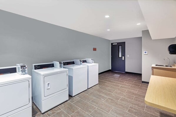 A laundry area with four white washers and dryers, a wooden counter with a sink, and a door leading to another room. - Tru by Hilton Laredo Airport (Laredo)