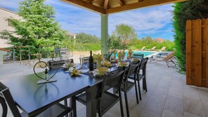 Une terrasse extérieure avec une table dressée pour le repas, entourée de chaises, et protégée par une structure de toit en bois.