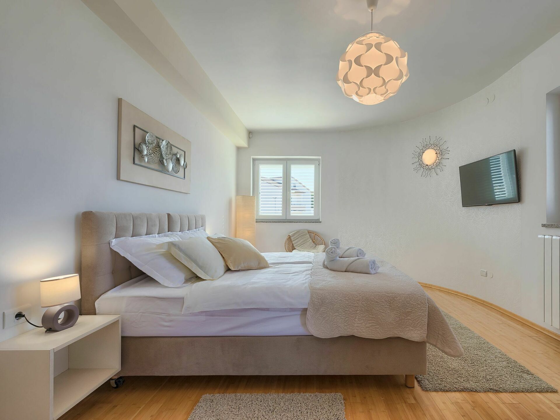 Un dormitorio moderno con una cama grande, una mesita de noche con lámpara, un televisor montado en la pared y una lámpara de techo original.